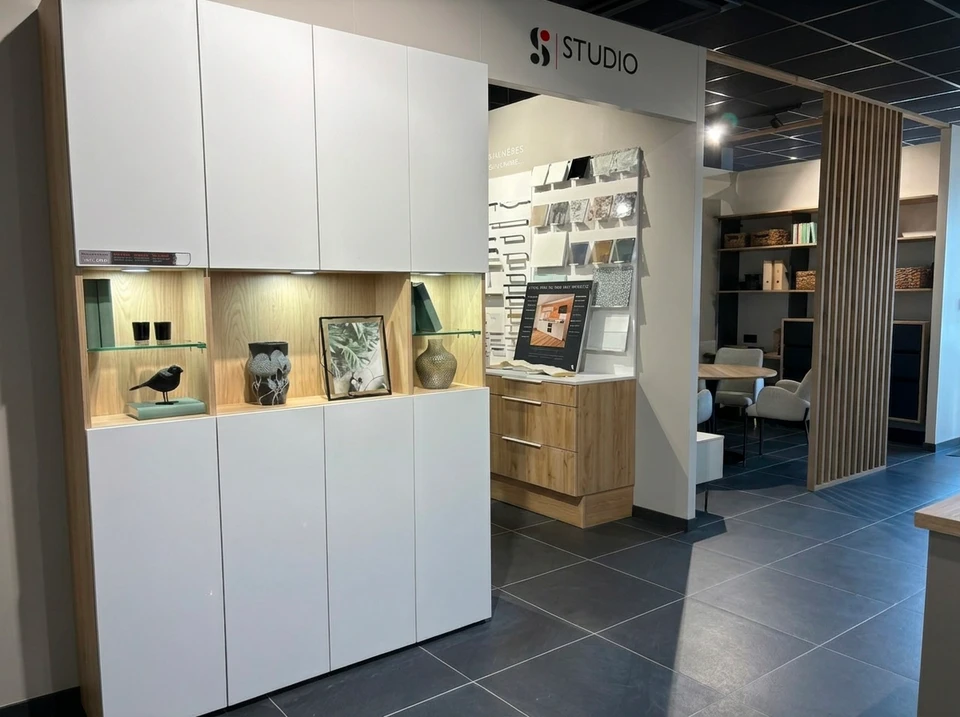 Fa&ccedil;ade moderne d'un showroom d'am&eacute;nagement int&eacute;rieur situ&eacute; dans une zone commerciale, avec une large devanture vitr&eacute;e illumin&eacute;e pr&eacute;sentant des am&eacute;nagements de cuisine et salle de bain en vitrine, parking accessible devant le b&acirc;timent, carrefour routier visible indiquant une bonne accessibilit&eacute; routi&egrave;re, clients se dirigeant vers l'entr&eacute;e du magasin en journ&eacute;e. Photo prise de face, mettant en avant la fa&ccedil;ade accueillante et la localisation dans une zone facilement accessible.